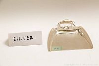 Front and side perspective of the sterling silver dust pan with a decorative handle and clean polished surface, shown alongside a handwritten 'SILVER' sign.