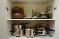 Wooden salad bowls stacked on the left shelf with metal graters, dome-shaped steamers, and cooling racks on the right shelf. Bottom shelf shows stainless steel cooking pots with lids and a non-electric pressure cooker with lid and handles.