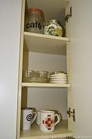 Photo shows shelves with cafe canister, floral ginger jar, glass juicer, small bowls or egg cups, and 3 coffee mugs on lower shelf.