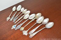 Photo showing all 11 sterling silver souvenir spoons laid out in a line on a brown surface. The spoons feature various detailed handles and bowls, some with colored enamel.