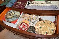 Open buffet drawer with assorted spoons and forks in utensil wrap, coasters, trivets, handmade tea cozy, Noritake vegetable dish with cover, paper plates, and other kitchen accessories.