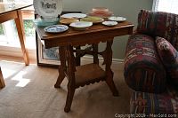 Wooden carved side table with various plates and bowls displayed on top, positioned beside a couch and near a window.