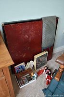 Red collapsible card table with visible wear marks leaning against the wall with items in front including cards, puzzle box, and dolls.