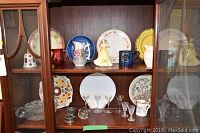 A wooden display cabinet with 7 decorative china plates in various patterns including floral and blue motifs, multiple pitchers in ceramic and glass, clear sherry glasses, and other small ceramic items.