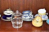 Teacups and saucers including Royal Chelsea dark blue and gold, Real Old Willow blue and white, yellow pottery items, and clear glass olive jar with spoon.