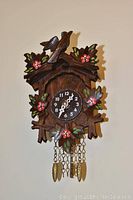 Front view of a wooden cuckoo clock decorated with carved birds and pink flowers, gold chains hanging.