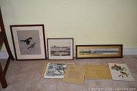 Shows three framed prints against a wall with a chair adjacent. Frames include a vertical bird illustration, a square cityscape, and a panoramic landscape. Below are several unframed prints and documents laid flat on the floor.