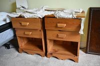Two matching wooden night stands side by side, showing their front faces with two drawers each and an open shelf below. The tops are covered partially by fabric.