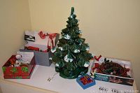 Photo showing electric ceramic Christmas tree with a power cord, small decorative boxes with ornaments, and a snowman figure on a shelf.