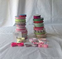 Photo shows 29 rolls of grosgrain ribbon stacked in two piles approximately 14 and 15 rolls each, plus 6 loose ribbons laid out in front. Ribbons display various pink, green, white, and red colors with different themed prints including polka dots, floral, hearts, and Easter motifs.