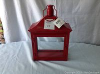 Red metal lantern with glass door and top handle, tags still attached indicating new condition, set against a white fabric background.