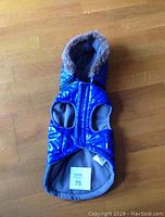 Blue waterproof winter dog coat with gray faux fur trimmed hood lying flat on wooden floor, size 75 tag visible.