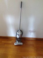 Shark stick vacuum cleaner upright against wall on wooden floor.