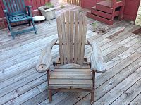 Front view of weathered wooden Adirondack chair with visible wear on armrests and seat, placed on wooden deck.