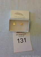Pair of yellow-toned sterling silver 'Love' shaped stud earrings inside white Magpie branded box on a white textured surface next to auction lot number 131.