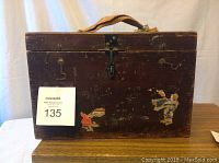 Photo of vintage wooden box showing front with metal latch lock, hooks, woven handle, and worn decals.