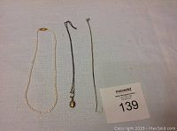 Photo showing three necklaces lying flat on a light-colored fabric background: a freshwater pearl necklace, a tiny silver locket on a black/dark chain, and a plain thin silver chain.