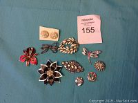 Photo of 11 vintage brooches and buttons arranged on a blue-green fabric background, showing a variety of floral and decorative designs including enamel, rhinestones, and metalwork.