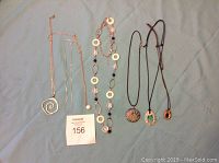 Six necklaces laid out on a light grey fabric background, showing various chain and cord styles with different pendants.