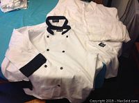 Set of three chefs jackets displayed on table; two plain white, one with black collar and buttons.