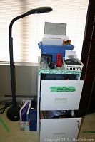 View of Verilux adjustable office floor lamp next to metal two-drawer filing cabinet filled with office supplies including trays, papers, and folders.