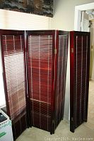 Two wood room dividers, each with 4 panels with adjustable wood blinds, shown folded and standing upright inside a room.