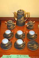 Full tea set arranged on wooden surface showing teapot, creamer, sugar bowl, 5 cups, 6 saucers with black and gold Asian scenic design.