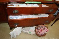 Opened wooden drawers with assorted white vintage linens inside and a wicker basket holding more linens on the floor.