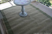 Full view of the outdoor carpet with a green border and beige geometric woven pattern. Shows placement of a small round table on the carpet to illustrate scale.