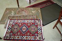 Five assorted rugs and mats stacked on carpeted floor, showing traditional oriental and floral patterns.