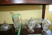 Full set of pressed glassware arranged on a table including jug, lidded dishes, butter dish, cake stand, and glasses. Shows overall collection and size.