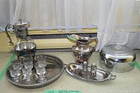 Full view of silver plate servingware including two pitchers on stands, six glass cups with silver plate detail on a large round tray, an oval tray with salt and pepper shakers, and a large domed lidded serving dish.
