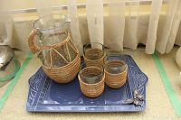 Pitcher with woven palm fibre handle and four matching glasses placed on a rectangular blue pewter tray with seagull design, next to window with curtains.