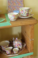 Royal Winton Chintz tea set pieces on wooden shelf with floral patterns, including teapot, creamer, sugar bowl, cups, and saucers alongside a floral-patterned book.