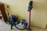 Photo showing Kenmore Elegance 360 canister vacuum cleaner with hose and wand, and Dyson V6 stick vacuum cleaner leaning against wall with basket of accessories.