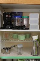 Magic Bullet single shot blender base with blade, three measuring cups, three plastic storage canisters with lids stacked on right, green ceramic ramekins, glass measuring cup, stainless steel measuring cup, plastic measuring cup with black markings, tall glass bottle with white lid and green handle