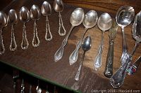 Top shelf view of assorted silver plated spoons showing various designs and sizes lined up horizontally on wood shelf.