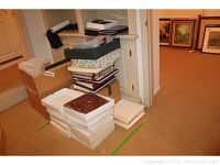Stack of photo albums in white storage boxes and some loose, located in corner next to picture frames and framed artwork on floor.