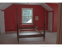 Four-poster wooden bed frame viewed from the side in an empty room with red walls and beige carpet.