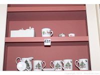 Photo showing shelves with pitcher and six mugs all decorated with Christmas tree holiday pattern, plus Alice in Wonderland mug and napkin rings.