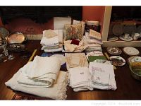 Wide shot showing stacks of white and off-white linens including tablecloths, napkins, needlepoint pillow, and doilies arranged on a table with some china plates to the side.