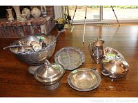 Photo of silverplate punch bowl holding punch cups and ladles, with other silverplate serving items around it on wooden floor.