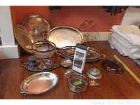 Wide view showing multiple silverplate serving platters, glass bowls, metal cheese grater, ashtray, trinket dish, and spoons arranged on a wooden surface.