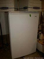 Front view of white Kenmore mini fridge, closed door, standing on garage floor with some boxes in background.