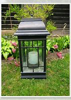 Full view of black metal framed solar garden lantern with glass panels and candle light inside, placed on grass with flowers in background.