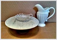 Set of three items side by side on cardboard background: marble pedestal plate, cut crystal bon bon dish, and white ceramic pitcher