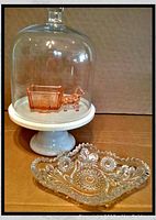 Glass dome pedestal cake stand with white base holding the pink depression glass horse/donkey cart candy dish and placed next to a clear cut crystal bon bon dish.