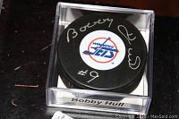 Top view of the black hockey puck showing the Winnipeg Jets logo and Bobby Hull's autograph in silver ink, inside clear plastic display case.