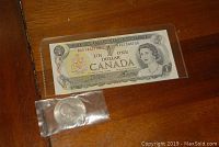 Uncirculated Canadian one dollar banknote with 1965 silver dollar coin in plastic sleeve, on wooden surface.