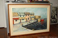 Framed oil on canvas depicting seaside dock with houses, boats, and birds. Shows full painting and frame on hardwood floor.